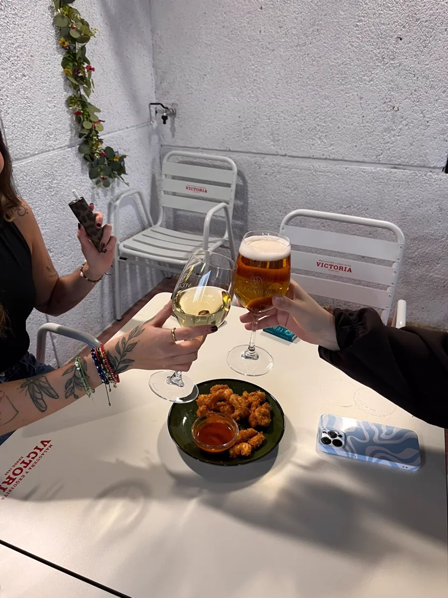 Dos clientas brindando con una copa de vino blanco y una caña, junto a una ración de pollo crujiente